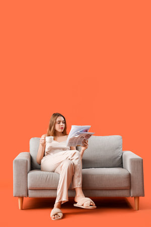 Young woman in pajamas with coffee cup reading newspaper on gray sofa against orange backgroundの写真素材