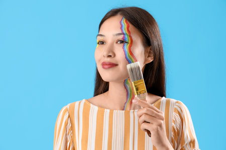 Young Asian woman with painted face and brush on blue background, closeup