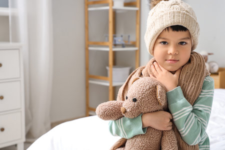 Ill little Asian boy with toy bear suffering from sore throat in bedroomの写真素材