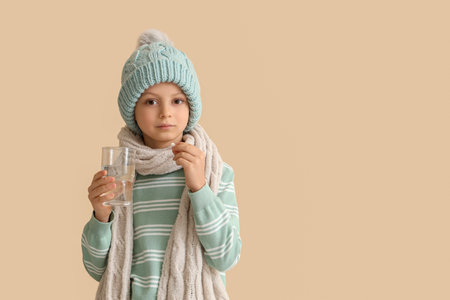 Ill little boy in hat with pill and glass of water on beige backgroundの写真素材