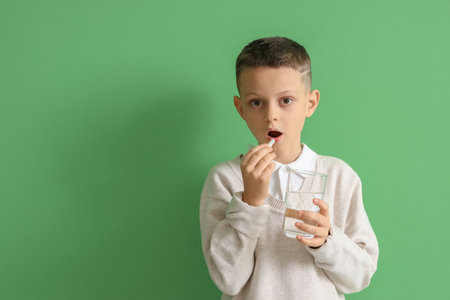 Ill little boy with glass of water taking pill on green backgroundの写真素材