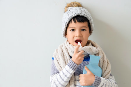 Ill little Asian boy with hot water bottle using spray for sore throat on light backgroundの写真素材