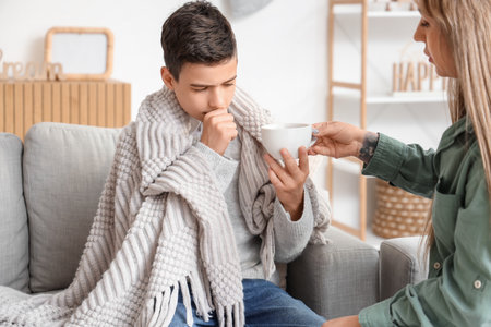 Sick little boy taking cup of tea from his mother at homeの写真素材