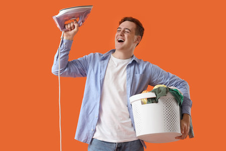 Cheerful young man with electric iron and full laundry basket on orange backgroundの写真素材
