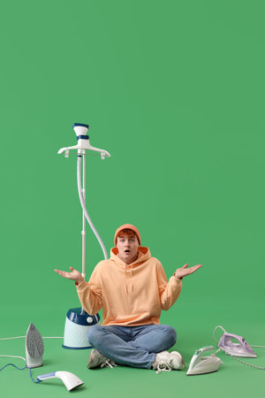 Confused young man with garment steamer and different electric irons on green background