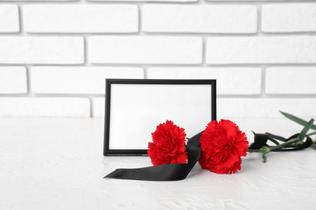 Blank funeral frame, carnation flowers and blank ribbon on table near light brick wallの写真素材