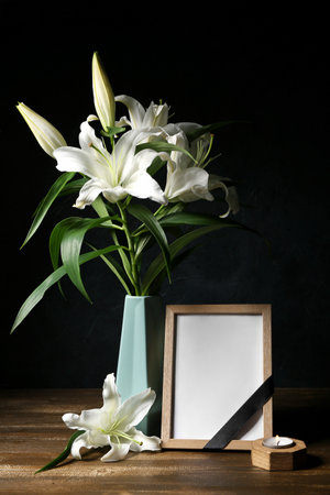 Blank funeral frame and vase with lily flowers on wooden table against dark backgroundの写真素材