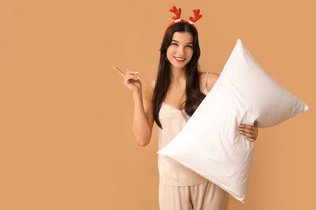 Beautiful young woman in silky pajamas and reindeer horns with soft pillow pointing at something on brown backgroundの写真素材