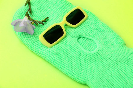 Stylish balaclava with flower and sunglasses on green background