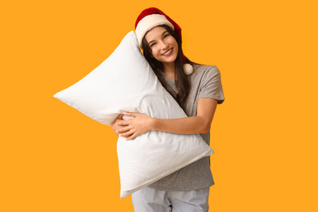 Beautiful young woman in pajamas and Santa hat with soft pillow on yellow backgroundの写真素材