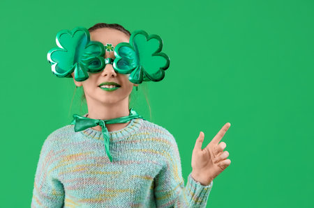 Funny girl with face painting and clover shaped novelty glasses pointing at something on green background. St. Patrick's Day celebrationの写真素材