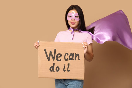 Young woman in superhero costume holding cardboard with text WE CAN DO IT on beige backgroundの写真素材