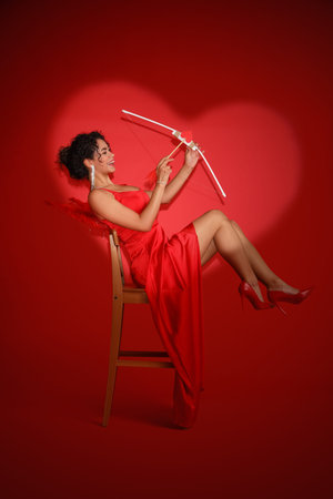 Young African-American woman dressed as Cupid with bow on red background. Valentine's Day...