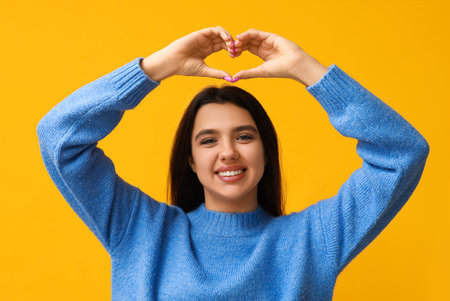 Happy young girl showing heart gesture on yellow background. Valentine's Day celebrationの写真素材
