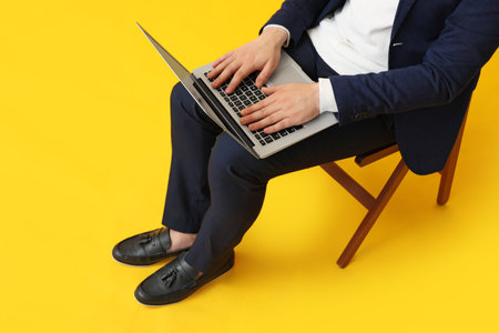 Young man in black stylish shoes with modern laptop sitting on chair against yellow backgroundの写真素材