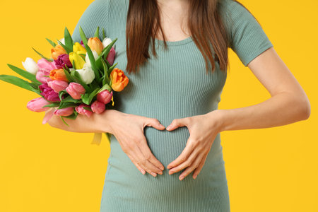 Young pregnant woman with bouquet of tulips showing heart gesture on yellow background....