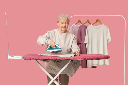 Senior woman with rack of clothes ironing laundry on pink backgroundの写真素材