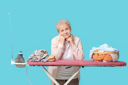 Displeased senior woman with clean clothes and ironing board on blue backgroundの写真素材