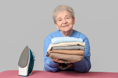 Senior woman with stack of laundry and iron on gray backgroundの写真素材
