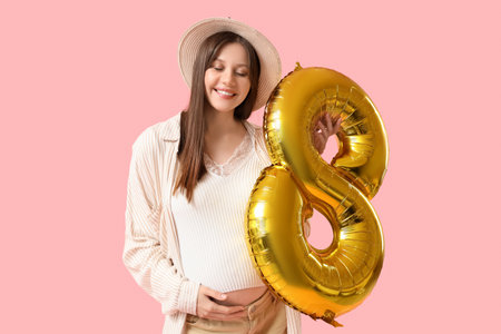 Beautiful young pregnant woman with golden air balloon shape of figure 8 on pink background. International Women's Dayの写真素材