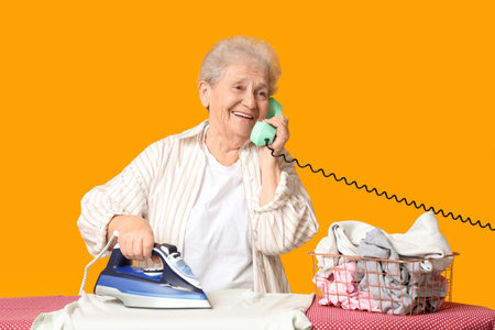 Happy senior woman ironing clothes and talking by phone on yellow backgroundの写真素材