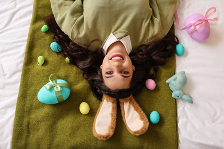 Young woman in bunny ears with Easter eggs lying on bed, top view