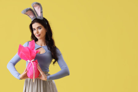 Young woman in bunny ears with Easter gift egg on yellow backgroundの写真素材