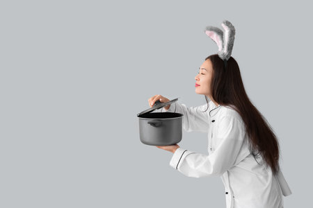 Female Asian chef in bunny ears with cooking pot on light background. Easter celebrationの写真素材