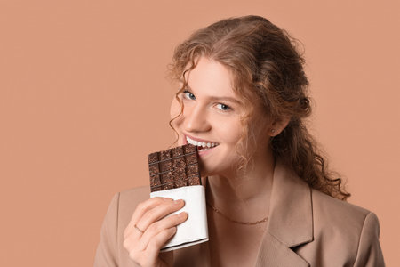 Beautiful young woman eating sweet chocolate bar on brown backgroundの写真素材
