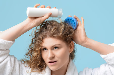 Beautiful young woman using hair scalp massager with bottle of shampoo on blue background