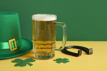 Mug of beer with paper clovers, leprechaun hat and horseshoe on yellow table against green background. St. Patrick's Day celebrationの写真素材