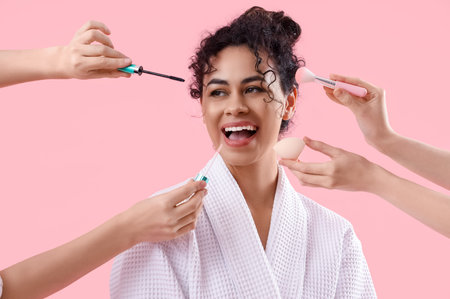 Young African-American woman in bathrobe and hands with makeup products on pink background, closeup