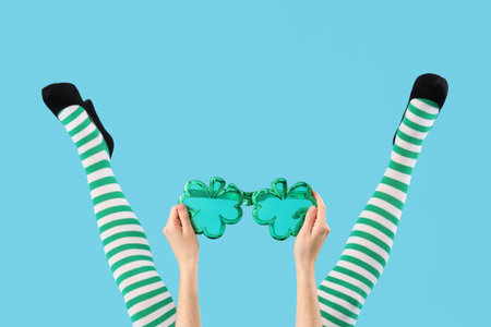 Female legs in green stockings with clover shaped novelty glasses on blue background. St. Patrick's Day celebrationの写真素材