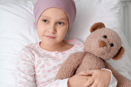 Little girl after chemotherapy with toy bear lying in bed at hospital, closeup. International Childhood Cancer Dayの写真素材