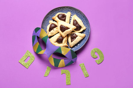 Plate with tasty Hamantaschen cookies, carnival mask and word PURIM on color background