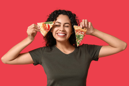 Beautiful young happy African-American woman with slices of delicious pizza on red backgroundの写真素材