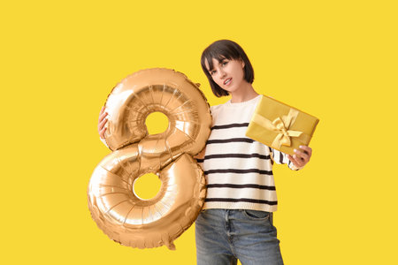 Beautiful young woman with balloon in shape of figure 8 and gift on yellow background. International Women's Dayの写真素材