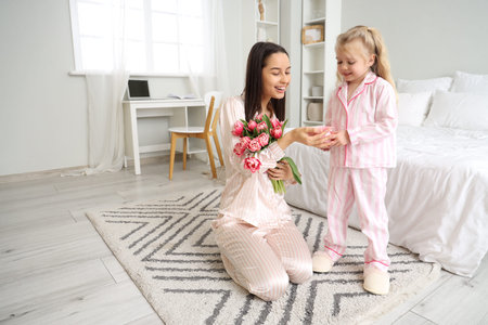 Little girl congratulating her happy mom with bouquet of beautiful flowers and gift box in bedroom. Mother's Day celebrationの写真素材