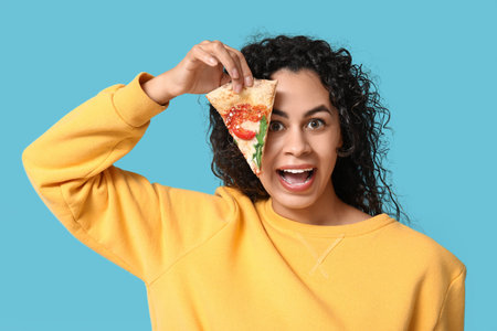 Beautiful young happy African-American woman with slice of delicious pizza on blue backgroundの写真素材