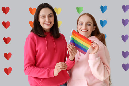 Young lesbian couple with LGBT flag against rainbow hearts on light background. Valentine's Day...