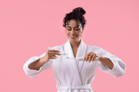 Young African-American woman in bathrobe squeezing tooth paste onto brush on pink backgroundの写真素材
