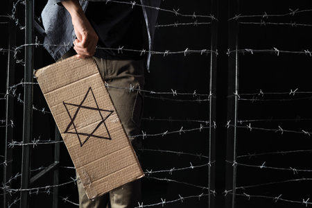 Young Jewish man holding paper with David star behind barbed wire on black background. International Holocaust Remembrance Dayの写真素材