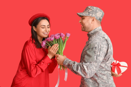 Man in military uniform greeting his wife with flowers and heart-shaped gift box on red background. Valentine's Day celebrationの写真素材