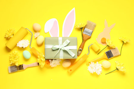 Painter's tools with Easter eggs, gift box and paper bunnies on yellow background