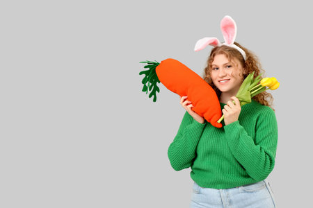 Beautiful young happy woman in bunny ears with carrot-shaped toy and tulip flower on gray...