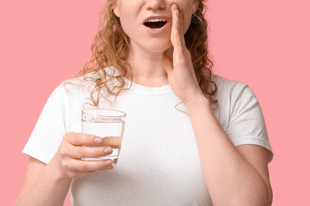 Beautiful young woman with glass of water suffering from toothache on pink background, closeup