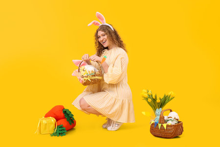 Beautiful young woman in bunny ears with Easter baskets, carrot-shaped toys and flowers on yellow...
