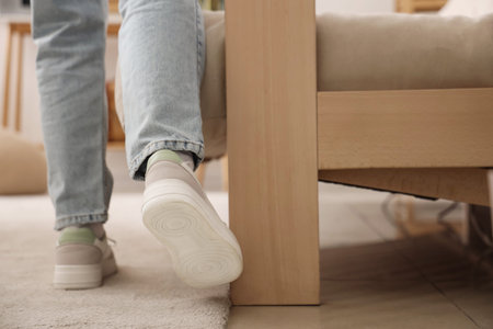 Young woman banging her foot on sofa at home, back view. Trauma concept
