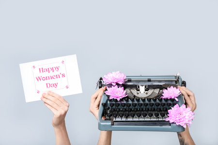 Hands holding vintage typewriter, flowers and festive postcard with text HAPPY WOMEN'S DAY on gray backgroundの写真素材