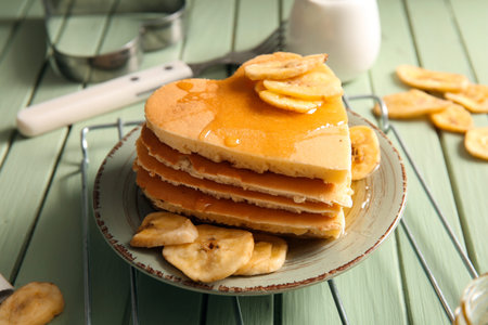 Plate with tasty pancakes in the shape of heart and dried banana on green wooden background,...